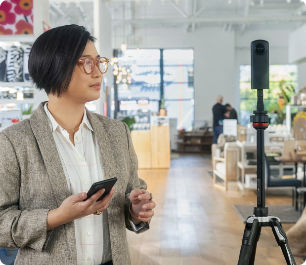 Woman standing in front of a 360 camera mounted on a tripod, holding her phone, in a modern office environment