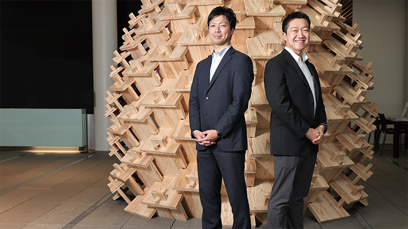 Two Japanese businessmen stand in front of an intricate wooden art installation
